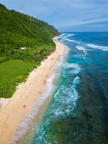 Aerial view of Pantai Nunggalan featuring a pristine sandy beach with turquoise waves crashing against the shore bordered by lush green cliffs in Kecamatan Kuta Selatan Bali Indonesia.