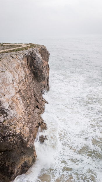 Aerial view of the rugged limestone cliffs of Sagres with white waves crashing against the rocky shore under an overcast sky Sagres Faro Portugal. Aerial view of the rugged limestone cliffs of Sagres with white waves crashing against the rocky shore under an overcast sky Sagres Faro Portugal.