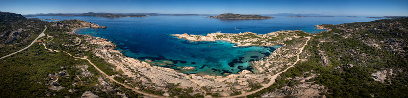 Aerial view of the rugged coastline of Carlotto with turquoise waters rocky cliffs and a winding coastal road under a clear blue sky in La Maddalena Sardinia Italy. Aerial view of the rugged coastline of Carlotto with turquoise waters rocky cliffs and a winding coastal road under a clear blue sky in La Maddalena Sardinia Italy.