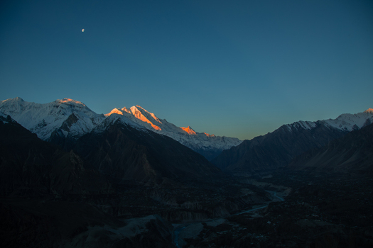 View of snow capped peaks of the Rakaposhi mountain range glowing under golden sunset light with a crescent moon in the clear sky Hunza Nagar Gilgit Pakistan. View of snow capped peaks of the Rakaposhi mountain range glowing under golden sunset light with a crescent moon in the clear sky Hunza Nagar Gilgit Pakistan.