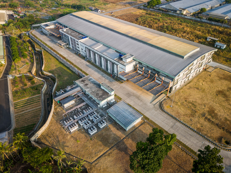 Aerial view of a large industrial warehouse facility with a metal roof loading docks and HVAC units surrounded by dry fields and vegetation in Semarang Regency Central Java Indonesia. Aerial view of a large industrial warehouse facility with a metal roof loading docks and HVAC units surrounded by dry fields and vegetation in Semarang Regency Central Java Indonesia.