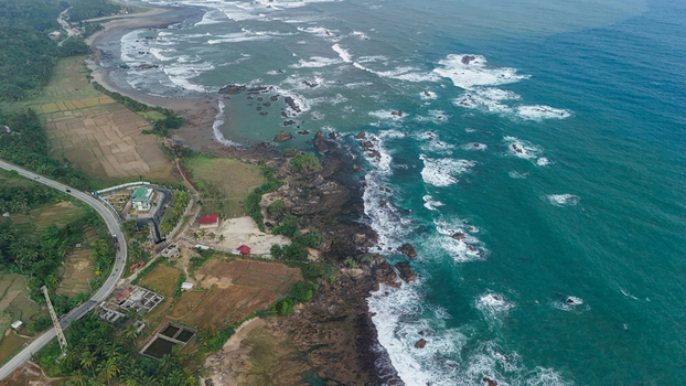 Aerial view of a rugged coastline with crashing waves a winding coastal road and agricultural fields in Indonesia. Aerial view of a rugged coastline with crashing waves a winding coastal road and agricultural fields in Indonesia.