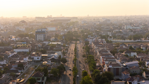 Indonesia 24 September 2026 Aerial view of a sprawling urban landscape with dense residential rooftops a central road lined with trees and a hazy horizon under a soft golden light.
