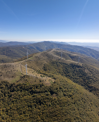 Aerial view of wind turbines situated on a mountain ridge surrounded by dense forests under a clear blue sky in Bulgaria. Aerial view of wind turbines situated on a mountain ridge surrounded by dense forests under a clear blue sky in Bulgaria.