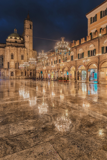 View of the historic Piazza del Popolo at night featuring the illuminated Cathedral of Sant Emidio ornate chandeliers and reflections on the wet pavement in Ascoli Piceno Marche Italy.
