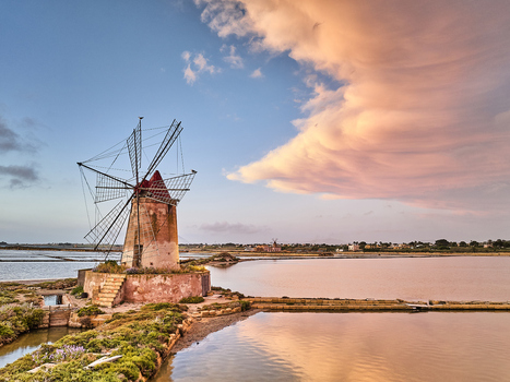 View of the historic windmill at Saline dello Stagnone with calm salt pans reflecting the pink sunset sky in Marsala Sicilia Italy.