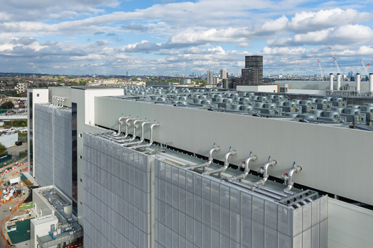 Aerial view of a large data center with industrial cooling units and pipes on the roof set against a city skyline under a cloudy sky in the United Kingdom. Aerial view of a large data center with industrial cooling units and pipes on the roof set against a city skyline under a cloudy sky in the United Kingdom.