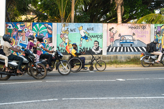 Sri Lanka 25 January 2026 View of motorcyclists and a person in a hand cycle passing a colorful mural wall along a roadside.
