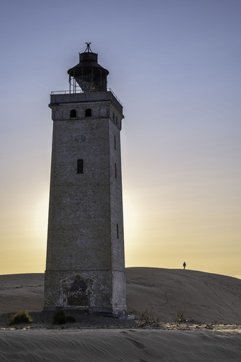 View of Rubjerg Knude lighthouse standing on sand dunes at sunset with a person walking in the distance in Lokken Denmark. View of Rubjerg Knude lighthouse standing on sand dunes at sunset with a person walking in the distance in Lokken Denmark.
