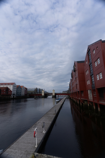 View of colorful wooden warehouses along the Nidelva river with a wooden pier and the Bakke Bridge under a cloudy sky in Trondheim Trondelag Norway. View of colorful wooden warehouses along the Nidelva river with a wooden pier and the Bakke Bridge under a cloudy sky in Trondheim Trondelag Norway.