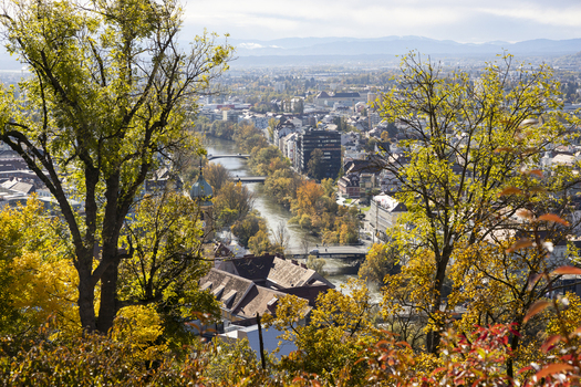 View of the Mur river snaking through the city with the autumn foliage painting the trees in fiery hues of red orange and gold Graz Styria Austria. View of the Mur river snaking through the city with the autumn foliage painting the trees in fiery hues of red orange and gold Graz Styria Austria.