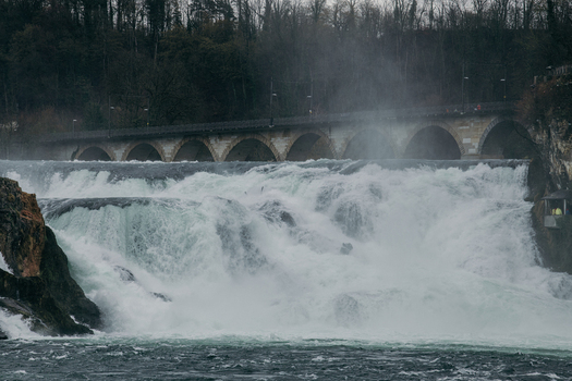 View of the Rhine Falls unleashing its raw power a torrent of white water contrasting with the dark mossy rocks and the arched bridge Schaffhausen Switzerland. View of the Rhine Falls unleashing its raw power a torrent of white water contrasting with the dark mossy rocks and the arched bridge Schaffhausen Switzerland.