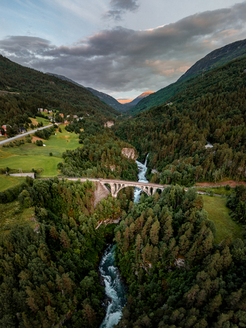 View of the historic Kylling Bridge spanning the Rauma river surrounded by lush green forests and dramatic mountains under a golden hour sky in Rauma Municipality More og Romsdal Norway. View of the historic Kylling Bridge spanning the Rauma river surrounded by lush green forests and dramatic mountains under a golden hour sky in Rauma Municipality More og Romsdal Norway.