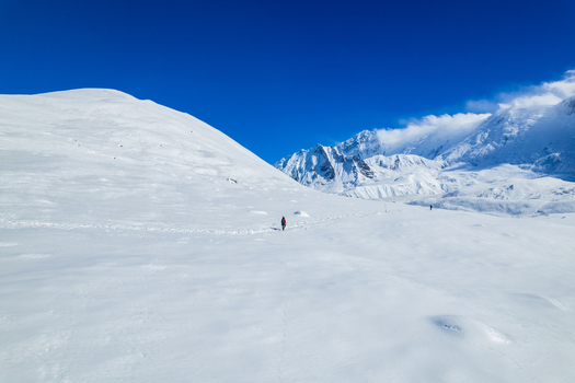 Aerial view of a lone hiker trekking across a vast snow covered mountain landscape under a clear blue sky in Nepal.