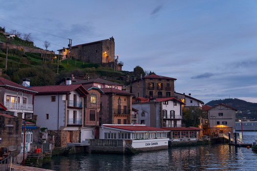 Pasai Donibane Spain 09 March 2026 View of the historic harbor street with traditional waterfront houses and a stone church on the hillside at dusk.