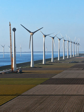 Aerial view of a row of modern wind turbines along a coastal dike with agricultural fields and a construction crane under a clear blue sky in Netherlands.