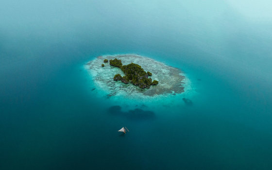 Aerial view of a small island with a dense green forest surrounded by turquoise waters a lone sailboat drifting nearby Pine Island L Île des Pins New Caledonia. Aerial view of a small island with a dense green forest surrounded by turquoise waters a lone sailboat drifting nearby Pine Island L Île des Pins New Caledonia.