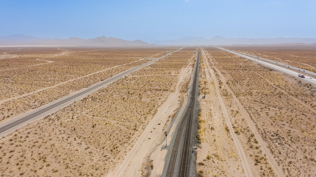 Aerial view of a highway and railway line running through the vast arid desert landscape under a clear blue sky in the United States.