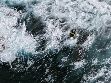 Aerial view of a surfer paddling through turbulent white foam and dark blue ocean waves at Praia do Areal de Santa Barbara Ribeira Grande Acores Portugal. Aerial view of a surfer paddling through turbulent white foam and dark blue ocean waves at Praia do Areal de Santa Barbara Ribeira Grande Acores Portugal.