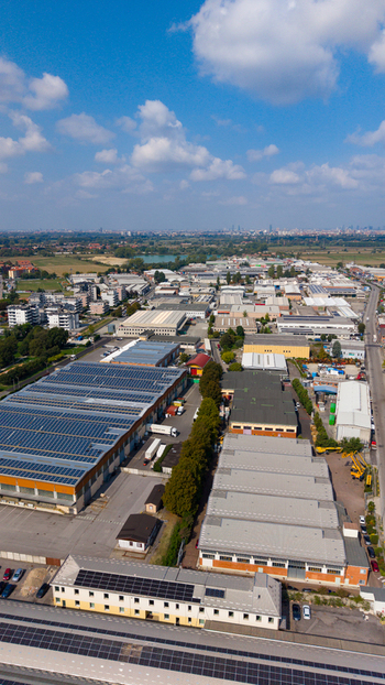 Aerial view of manufacturing and logistics buildings featuring extensive solar panel installations on rooftops surrounded by paved yards and green trees in San Giuliano Milanese Lombardy Italy.
