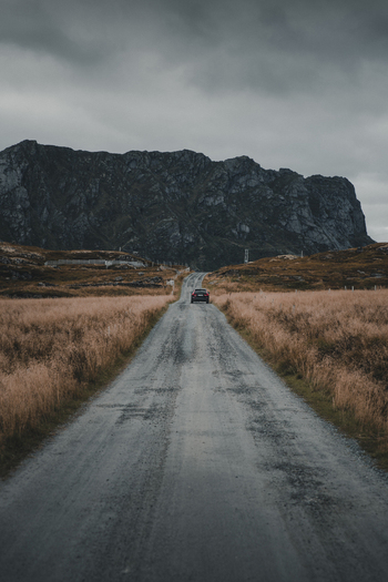 View of a car driving along a gravel road towards the rugged mountains of Nesland surrounded by dry grass under a dramatic sky in Flakstad Municipality Nordland Norway. View of a car driving along a gravel road towards the rugged mountains of Nesland surrounded by dry grass under a dramatic sky in Flakstad Municipality Nordland Norway.