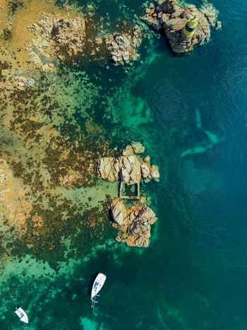 Aerial view of the lighthouse and rocky coastline with clear turquoise waters and small boats in Ile de Brehat Brittany France.