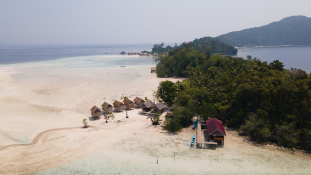 Aerial view of Ransiwor homestay with wooden bungalows on a white sand beach surrounded by lush tropical vegetation and turquoise waters in Raja Ampat Southwest Papua Indonesia.