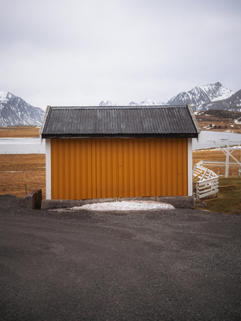 View of a mustard yellow building with a dark roof stands out against the snowy mountains and brown fields a stark contrast in the tranquil landscape Fredvang Nordland Norway. View of a mustard yellow building with a dark roof stands out against the snowy mountains and brown fields a stark contrast in the tranquil landscape Fredvang Nordland Norway.