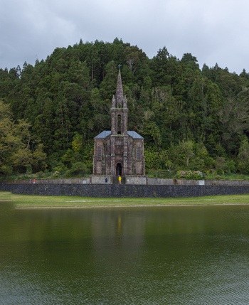 Aerial view of Ermida de Nossa Senhora das Vitorias situated by the lake surrounded by lush forest with people standing near the stone walls in Furnas Acores Portugal. Aerial view of Ermida de Nossa Senhora das Vitorias situated by the lake surrounded by lush forest with people standing near the stone walls in Furnas Acores Portugal.