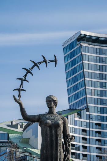 View of the First Swallows monument featuring a woman holding birds in flight against a modern glass skyscraper in Vilnius Vilnius County Lithuania. View of the First Swallows monument featuring a woman holding birds in flight against a modern glass skyscraper in Vilnius Vilnius County Lithuania.