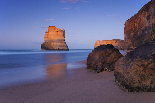 View of the Twelve Apostles limestone stacks from a sandy beach with large boulders and towering cliffs under a twilight sky in Australia. View of the Twelve Apostles limestone stacks from a sandy beach with large boulders and towering cliffs under a twilight sky in Australia.