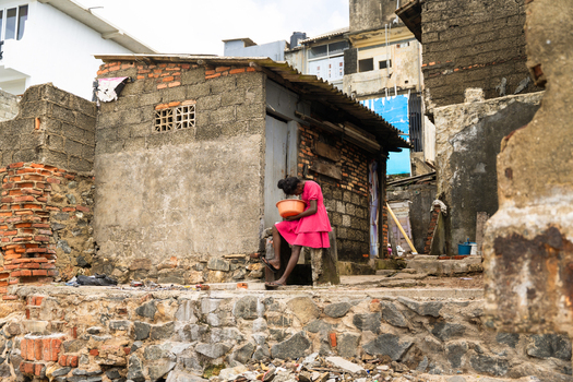 Sri Lanka 05 February 2026 View of a young girl in a pink dress sitting outside a weathered brick building holding an orange bowl in a poor urban area.