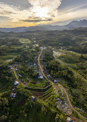 Aerial view of Bangka Kenda village surrounded by lush green hills terraced rice fields and a winding road under a dramatic sunset sky in Wae Rii East Nusa Tenggara Indonesia.