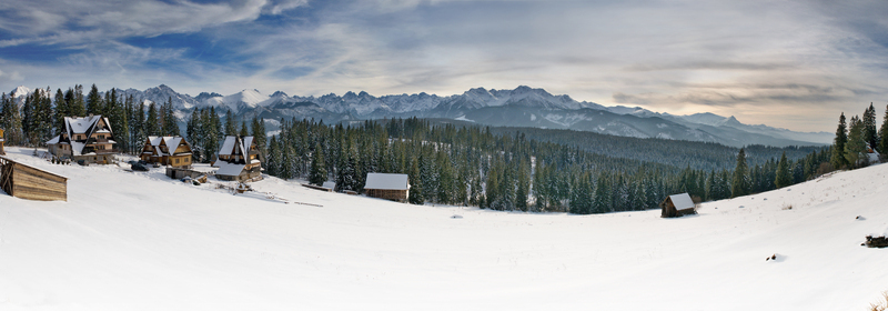 View of traditional wooden chalets on a snow covered field with the jagged peaks of the Tatra Mountains under a cloudy sky in Tatra County Lesser Poland Voivode Poland. View of traditional wooden chalets on a snow covered field with the jagged peaks of the Tatra Mountains under a cloudy sky in Tatra County Lesser Poland Voivode Poland.