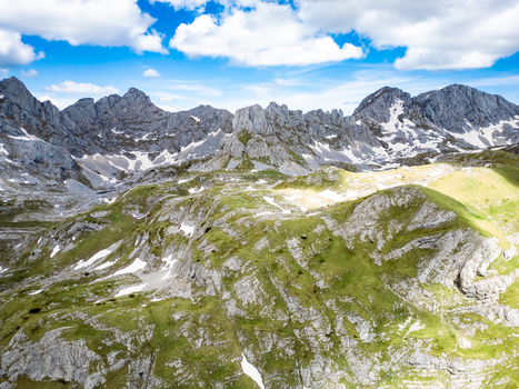 Aerial view of Bobotov Kuk in the Durmitor mountains featuring rugged rocky peaks patches of snow and green slopes under a bright blue sky in Zabljak Municipality Montenegro. Aerial view of Bobotov Kuk in the Durmitor mountains featuring rugged rocky peaks patches of snow and green slopes under a bright blue sky in Zabljak Municipality Montenegro.