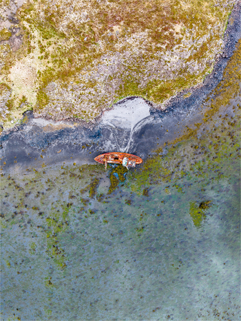 Aerial view of a rusted shipwreck resting on a dark volcanic beach surrounded by shallow turquoise waters and mossy green terrain in Iceland. Aerial view of a rusted shipwreck resting on a dark volcanic beach surrounded by shallow turquoise waters and mossy green terrain in Iceland.