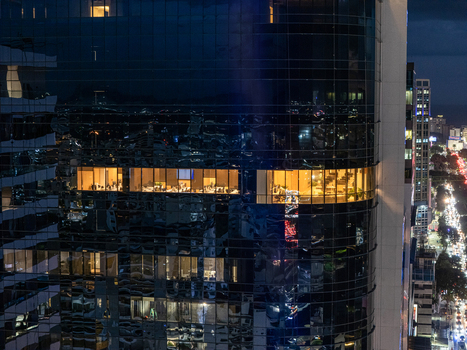 Aerial view of a modern glass skyscraper at night with glowing office interiors and city lights in the background in Santo Domingo Distrito Nacional Dominican Republic. Aerial view of a modern glass skyscraper at night with glowing office interiors and city lights in the background in Santo Domingo Distrito Nacional Dominican Republic.