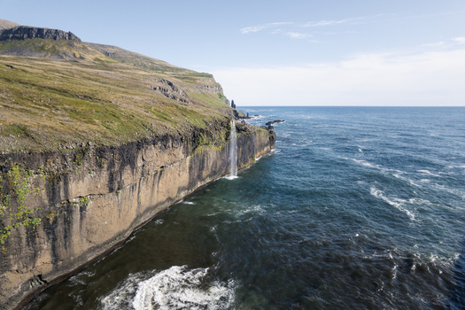 Aerial view of a waterfall cascading from the steep rocky cliffs of Hornstrandir Nature Reserve into the blue ocean Isafjordur Isafjardarbaer Iceland. Aerial view of a waterfall cascading from the steep rocky cliffs of Hornstrandir Nature Reserve into the blue ocean Isafjordur Isafjardarbaer Iceland.