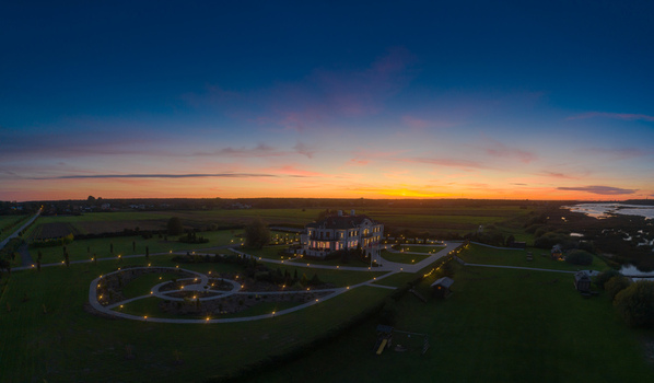 Aerial view of a grand mansion surrounded by manicured gardens and illuminated pathways during a vibrant sunset in Latvia. Aerial view of a grand mansion surrounded by manicured gardens and illuminated pathways during a vibrant sunset in Latvia.