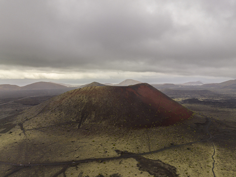 Aerial view of a striking red and brown volcanic crater surrounded by vast lava fields and rolling hills under a dramatic cloudy sky in Lanzarote Canarias Spain. Aerial view of a striking red and brown volcanic crater surrounded by vast lava fields and rolling hills under a dramatic cloudy sky in Lanzarote Canarias Spain.