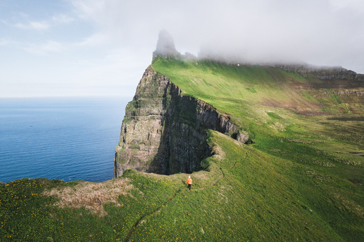 Aerial view of a hiker in an orange jacket walking along the dramatic green cliffs of Hornstrandir Nature Reserve overlooking the blue ocean Isafjordur Isafjardarbaer Iceland. Aerial view of a hiker in an orange jacket walking along the dramatic green cliffs of Hornstrandir Nature Reserve overlooking the blue ocean Isafjordur Isafjardarbaer Iceland.