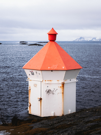 View of a white rust stained structure with a vivid orange roof stands resilient against the backdrop of the moody ocean and distant mountains Henningsv&aelig;r Norway.