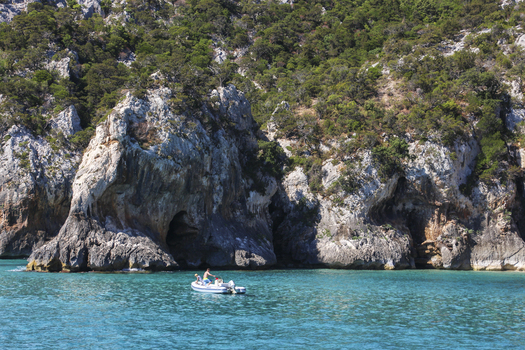 View of a small motorboat with people cruising in the turquoise waters of the Gulf of Orosei framed by rugged limestone cliffs and Mediterranean vegetation Italy. View of a small motorboat with people cruising in the turquoise waters of the Gulf of Orosei framed by rugged limestone cliffs and Mediterranean vegetation Italy.