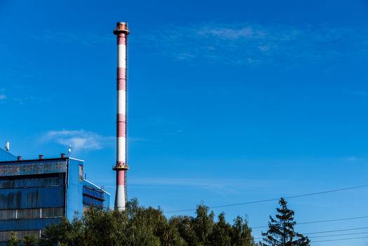 View of a tall red and white striped industrial chimney rising above a blue factory building and green trees under a clear blue sky in Poland.