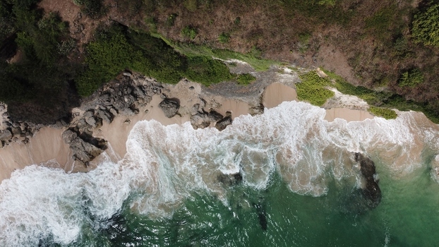 Aerial view of turquoise waves crashing against rocky cliffs and a sandy beach in Indonesia.