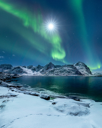 Aerial view of the aurora borealis dancing over snow capped mountains and a calm fjord under a bright moonlit sky in Nordland Norway.