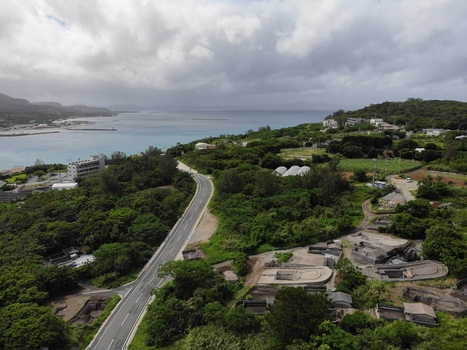 Aerial view of a winding asphalt road cutting through lush green vegetation on Sesoko Island with the Motobu Peninsula visible across the sea under a cloudy sky in Motobu Okinawa Japan. Aerial view of a winding asphalt road cutting through lush green vegetation on Sesoko Island with the Motobu Peninsula visible across the sea under a cloudy sky in Motobu Okinawa Japan.