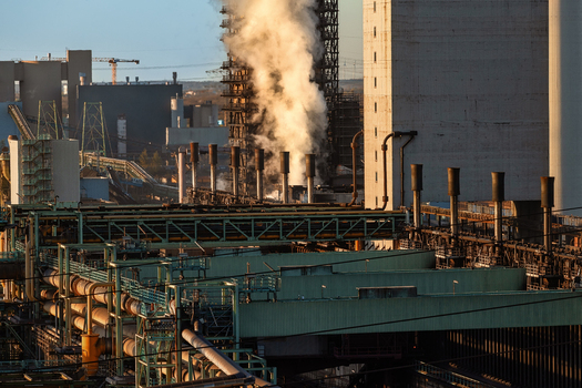 View of a massive industrial steel plant with thick white smoke rising from chimneys and a complex network of pipes in Duisburg North Rhine Westphalia Germany.