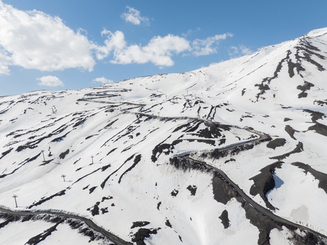 Aerial view of the snow covered south slope of Mount Etna with a winding road and ski lift pylons under a blue sky in Nicolosi Sicilia Italy. Aerial view of the snow covered south slope of Mount Etna with a winding road and ski lift pylons under a blue sky in Nicolosi Sicilia Italy.