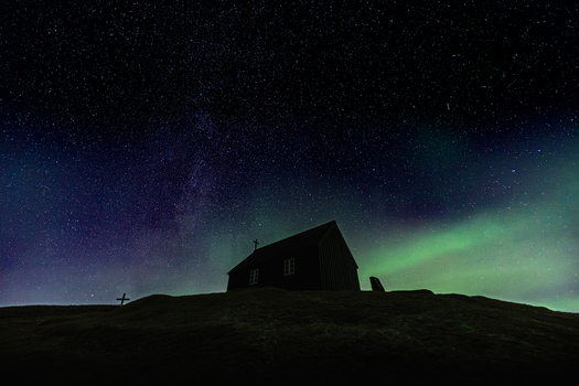 View of a lone dark church silhouette against a vibrant celestial canvas painted with the ethereal dance of the aurora borealis Krysuvikurkirkja Iceland.